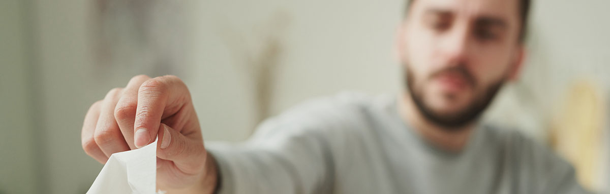 Hand of young sick businessman taking paper tissue from box while sitting by table in home environment and making working plan